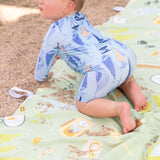A baby in a blue patterned outfit crawls on a Bumkins Reversible Roll & Go Blanket, Happy Camper, surrounded by sand on a sunny day.