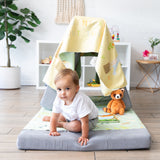 A toddler crawls under a gray cushion tent covered by a Bumkins Reversible Roll & Go Blanket, Happy Camper, with a plush toy nearby.