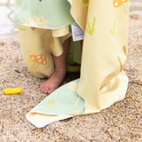 Close-up of a child’s bare feet peeking from below a Bumkins Reversible Roll & Go Blanket, Happy Camper on a sandy beach.