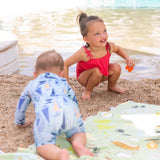 Two toddlers play on a beach, one in a red swimsuit with a toy and the other crawling on a Bumkins Reversible Roll & Go Blanket, Happy Camper.
