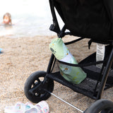Close-up of a stroller on the beach with a rolled Bumkins Reversible Roll & Go Blanket, Happy Camper in the basket, with sandals nearby.