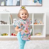 A toddler in a colorful Bumkins Ruffle Apron Bib: Boots and blue pants smiles in a playroom with toy-filled shelves behind.