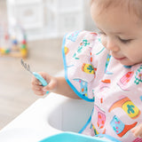 Close-up of a child in a high chair wearing a Bumkins Ruffle Apron Bib: Boots holding a fork, with blurred toys in the background.