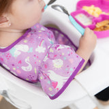 Close-up of a toddler in a high chair holding a blue spoon wearing a Bumkins Ruffle Apron Bib: Unicorns, with focus on the ruffle sleeve.