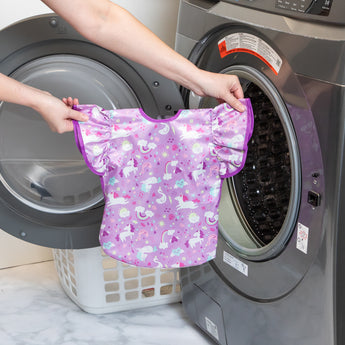 A person holds a bib from Bumkins Snack In Style 3-Piece Set: Unicorns and Rainbows in front of an open front-loading washing machine.