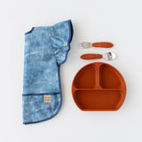 Bumkins Ruffle Apron Bib: Crushed Denim lays folded lengthwise on a white background next to an orange plate and matching utensils.