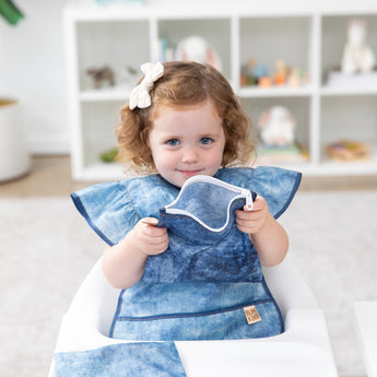 A young girl in a bow looks curiously at the camera with Bumkins Snack In Style 3-Piece Set: Denim On Denim.