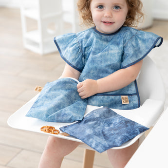 A girl smiles in a high chair with Bumkins Snack In Style 3-Piece Set: Denim On Denim.