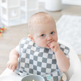 A baby in a Bumkins SuperBib® 3 Pack: Palms + Check sits in a high chair, nibbling curiously with a bowl in front of them.