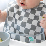 A baby in a checkered Bumkins SuperBib® 3 Pack: Palms + Check enjoys food in a high chair.