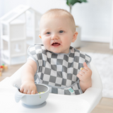 A baby sitting in a high chair wearing a checkered bib reaches into a bowl from Bumkins Baby-Led Weaning Essential 6-Piece Set: Gray.