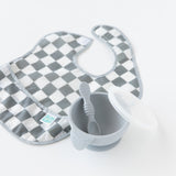 A gray Bumkins SuperBib® 3 Pack: Palms + Check sits gracefully beside a gray bowl with a spoon and lid on a pristine white background.