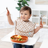 A smiling toddler in a Bumkins SuperBib® 3 Pack: Palms + Check sits in a high chair, holding utensils with food in a divided plate on the tray.