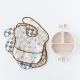 Bumkins SuperBib® 3 Pack: Palms + Check, made from waterproof fabric, next to a divided plate and utensils on a white background.