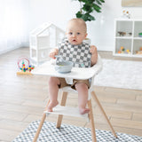 A baby in a high chair, wearing a Bumkins SuperBib® 3 Pack: Palms + Check, holding a spoon with a bowl on the tray.