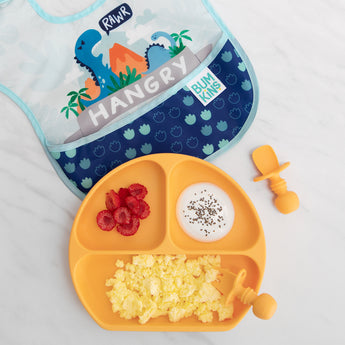 An orange plate with eggs, berries, and yogurt sits near a Bumkins SuperBib® 3 Pack: Hangry, Dinosaurs, Blue Tropic on a marble surface.