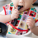 A child in a bright Bumkins SuperBib® 3 Pack: Mickey & Friends MVP holds a spoon, ready to eat from a bowl.
