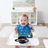 A smiling baby in a high chair, wearing a blue Bumkins SuperBib®: Mickey & Friends MVP, holds a spoon while eating cereal in a bowl.