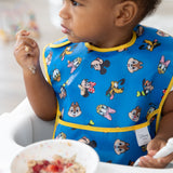 A toddler wears a blue Bumkins SuperBib® 3 Pack: Mickey & Friends MVP, holding cereal, with a bowl of fruit in front.
