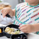 A toddler enjoys pasta and berries with a spoon from a divided tray, wearing a colorful Bumkins SuperBib® 3 Pack: Mickey & Friends MVP.