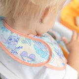 A toddler in a Bumkins SuperBib® 3 Pack: Stitch Star-ring Up Trouble looks down as they sit near an orange tray.