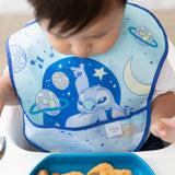 A child, sitting in a high chair, wears Bumkins SuperBib® 3 Pack: Stitch Star-ring Up Trouble and looks down at food on a blue plate.