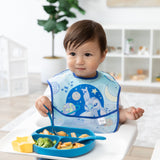 A toddler in a Bumkins SuperBib® 3 Pack: Stitch Star-ring Up Trouble munches from a divided blue plate in a bright, cozy room.
