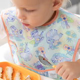 A toddler in a Bumkins SuperBib® 3 Pack: Stitch Star-ring Up Trouble, focused on eating from an orange plate with a spoon.