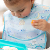 A toddler in a Bumkins SuperBib® 3 Pack: Stitch Star-ring Up Trouble holds orange food while looking down at a blue tray.