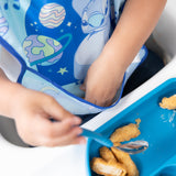 A child reaches into the pocket of a Bumkins SuperBib® 3 Pack: Stitch Star-ring Up Trouble while holding a spoon near nuggets on a blue plate.