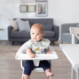 A baby sits in a high chair in a cozy living room, wearing a bib from Bumkins On-The-Go Mealtime 7-Piece Set: Pooh Bear + Friends.
