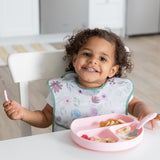 A girl in a floral bib smiles at a white table, eating breakfast with Bumkins Grip Dish Essential Toddler 4-Piece Mealtime Set: Pink.