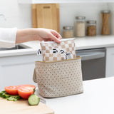A hand places Roamio by Bumkins Reusable Snack Bag 4 Pack: Double Check in a beige lunch bag on a kitchen counter next to sliced veggies.