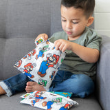 A young child eagerly opens Bumkins Reusable Snack Bag, Large: Super Mario™ & Luigi while sitting on a couch with other similar bags nearby.