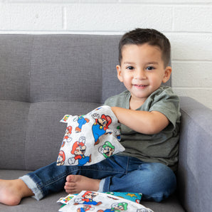 A smiling child sits on a gray couch, reaching into a Bumkins Reusable Snack Bag, Large: Super Mario™ & Luigi.