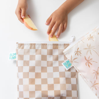 Close-up of a child placing apple slices into a Bumkins Reusable Snack Bag, Large 2 Pack: Palm Check on a white table with another bag nearby.