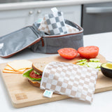 A kitchen scene with a wooden cutting board holding a sandwich in a checkered reusable bag, slices of tomato, cucumber, cheese, and half an avocado. A gray lunch bag is in the background on a white countertop.
