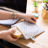 Hands grab a sandwich from a white Roamio by Bumkins Reusable Snack Bag, Large 2 Pack: Rich & Boujee on a desk next to a laptop.