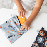 A child puts a smiley face cookie in a Bumkins Reusable Snack Bag, Large 2 Pack: Everyday Is Halloween at a white table.