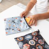 A child places a cookie into a Bumkins Reusable Snack Bag, Large 2 Pack: Everyday Is Halloween on a white table.