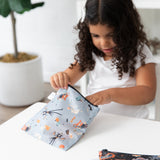 A young girl with curly hair opens a Bumkins Reusable Snack Bag, Large 2 Pack: Everyday Is Halloween at a table, a plant in the background.
