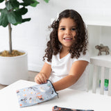 A curly-haired girl at a table reaches into a Bumkins Reusable Snack Bag, Large 2 Pack: Everyday Is Halloween, with a plant and shelves nearby.