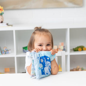 A smiling child holds Bumkins Reusable Snack Bag, Large 2 Pack: Stitch Star-ring Up Trouble at a table in front of toys on white shelves.