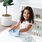 A child with curly hair reaches into a Bumkins Reusable Snack Bag, Large 2 Pack: Stitch Star-ring Up Trouble at a white table.