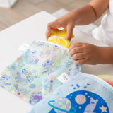 A child puts a yellow cookie in a Bumkins Reusable Snack Bag, Large 2 Pack: Stitch Star-ring Up Trouble on a white table.