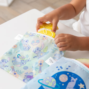 A child puts a yellow cookie in a Bumkins Reusable Snack Bag, Large 2 Pack: Stitch Star-ring Up Trouble on a white table.