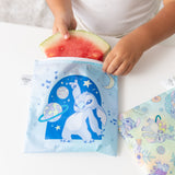 A child puts a watermelon slice into a Bumkins Reusable Snack Bag, Large 2 Pack: Stitch Star-ring Up Trouble on a white table.