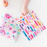 A child's hand reaches into bags from Bumkins Lunchbox Legends 6-Piece Set: Watercolor Pink Jelly on a white table.
