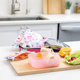 A kitchen counter with Bumkins Lunchbox Legends 6-Piece Set: Watercolor Pink Jelly, lunch bag, cutting board, and fruit & veggies.