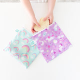 A child places a heart-shaped sandwich in bags from Bumkins Lunchbox Legends 6-Piece Set: Unicorns Purple Jelly at a white table.
